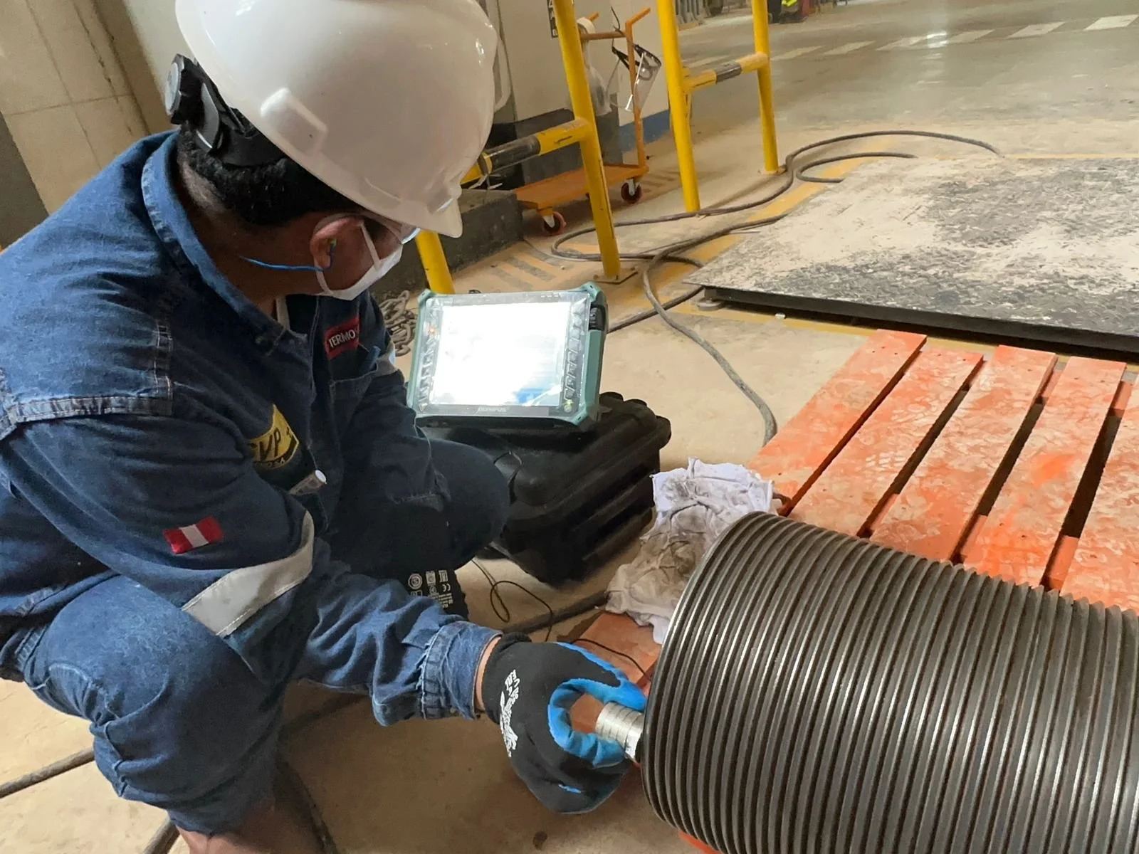 Trabajador con casco, mascarilla, guantes y uniforme industrial inspecciona un componente metálico grande en un entorno de construcción o mantenimiento.