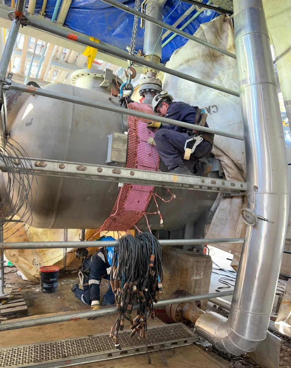 Dos trabajadores en equipo de construcción inspeccionando una gran estructura metálica en un sitio industrial.