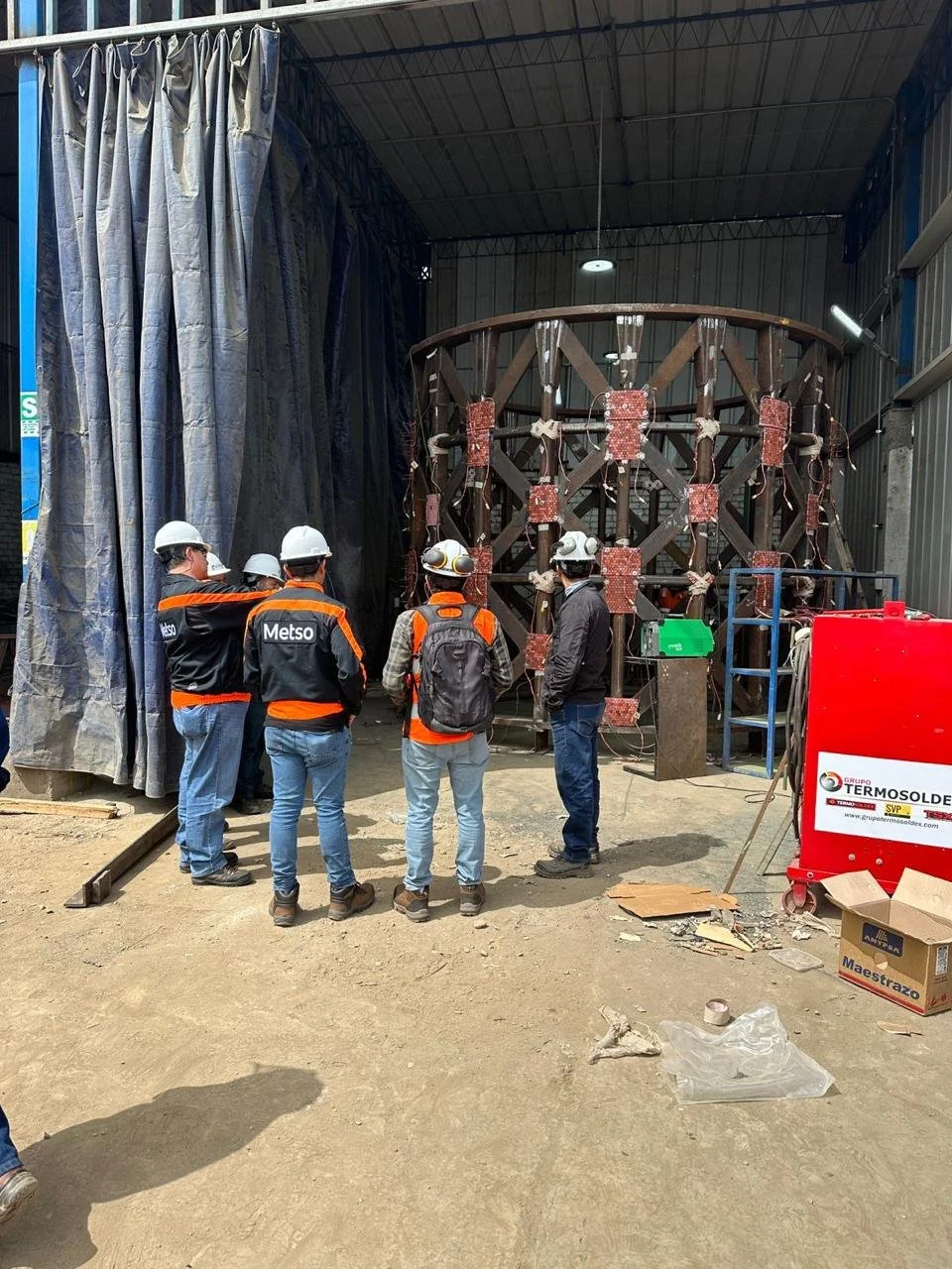 Grupo de trabajadores con cascos y chalecos de seguridad observando una estructura metálica en construcción en un espacio cerrado con paredes de metal y cortinas azules.