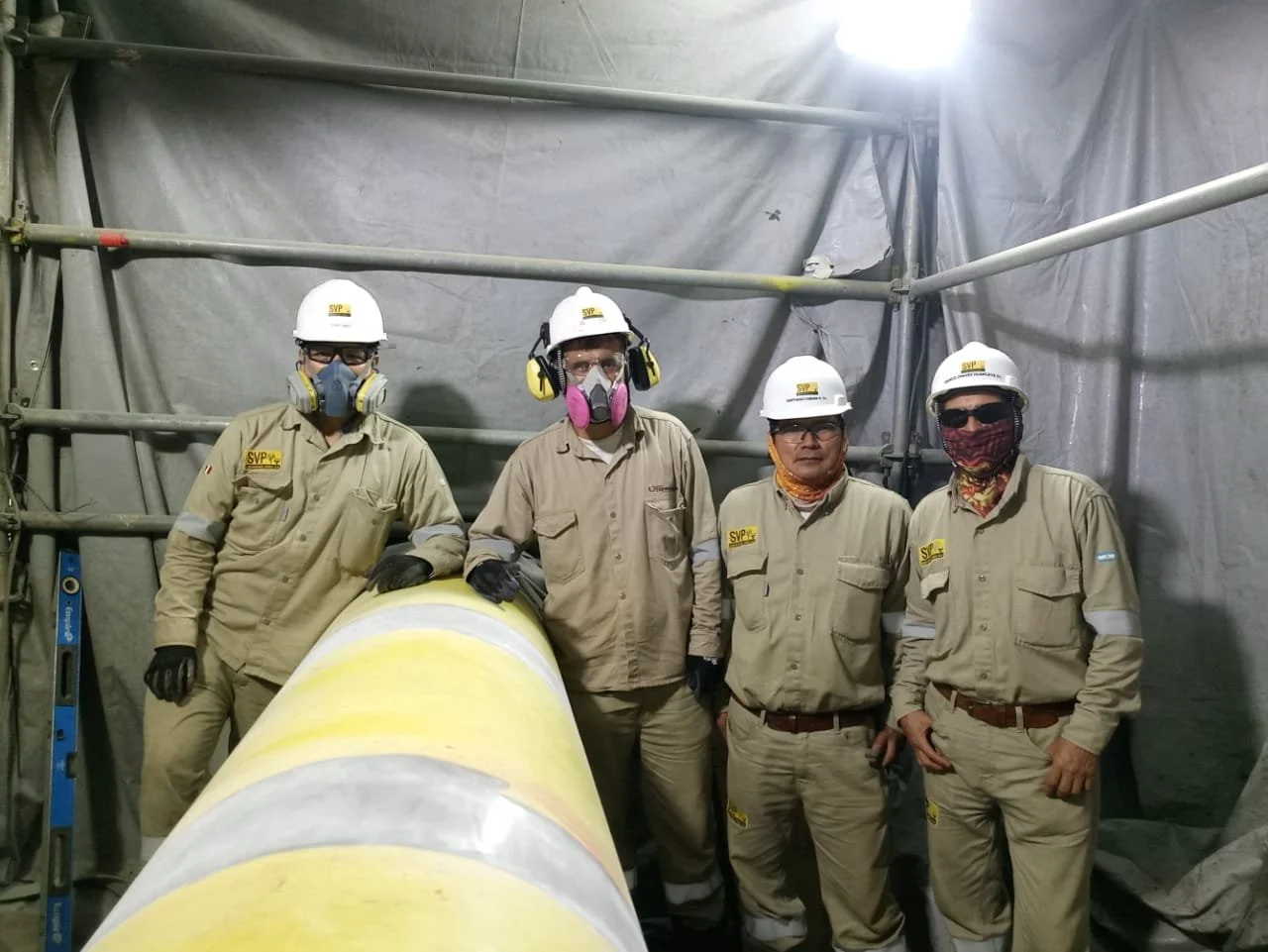 Cuatro empleados con casco y equipo de protección en un ambiente industrial, junto a un tubo grande de color amarillo con gris.