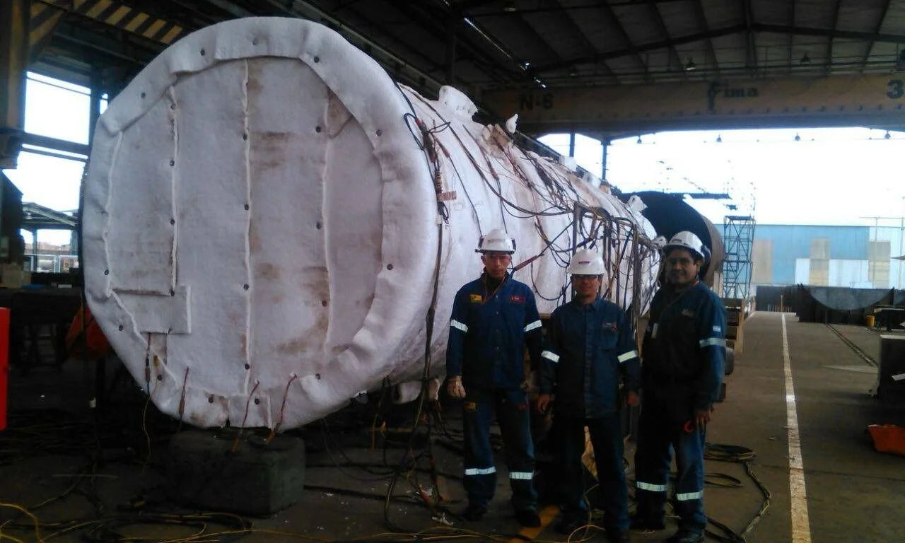 Tres trabajadores con casco y uniformes de seguridad posando frente a una gran estructura industrial en un taller o fábrica.