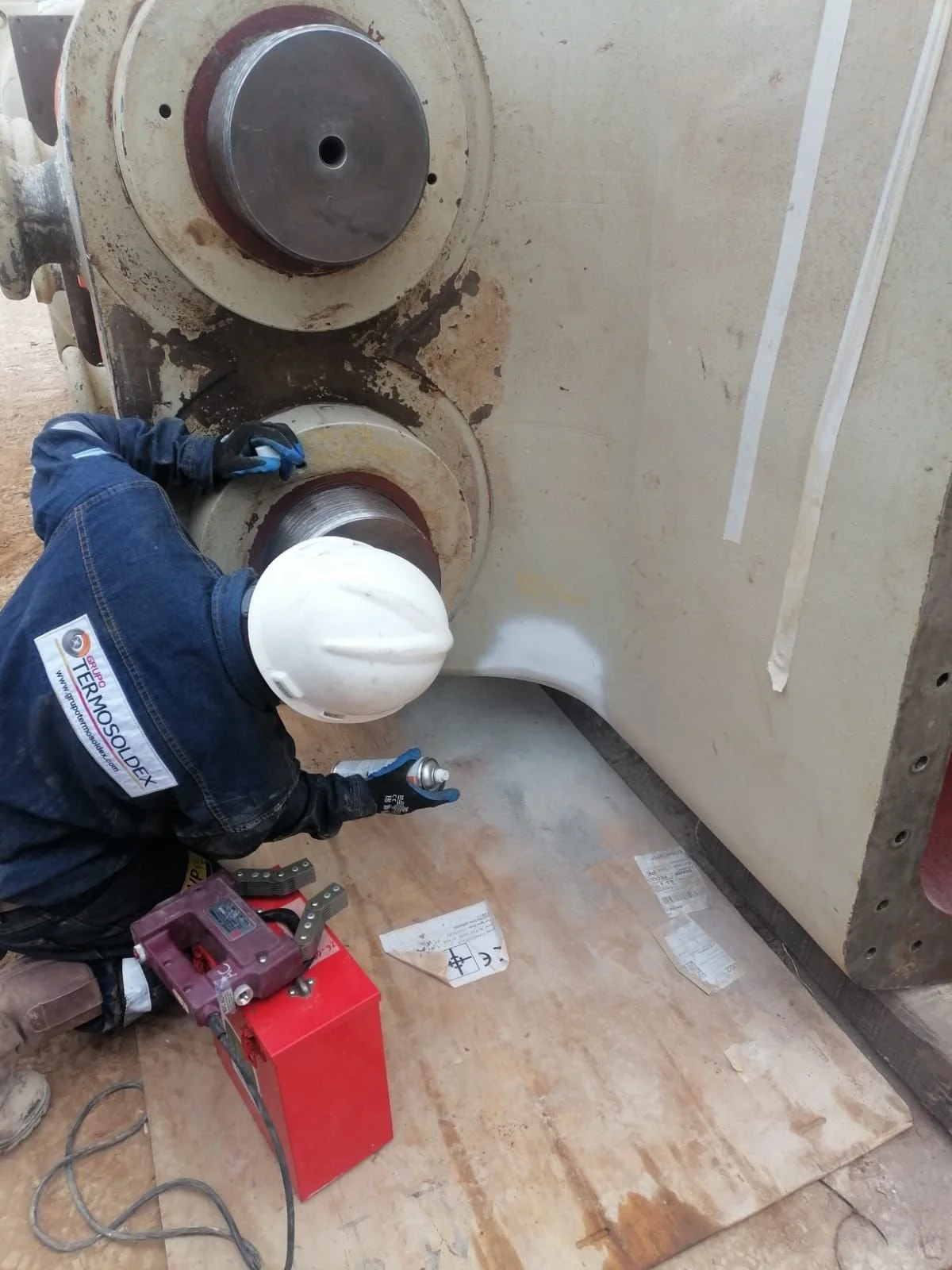 Trabajador usando equipo de protección mientras trabaja en una gran pieza de maquinaria industrial, aplicando pintura o anticorrosivo con un aerosol.
