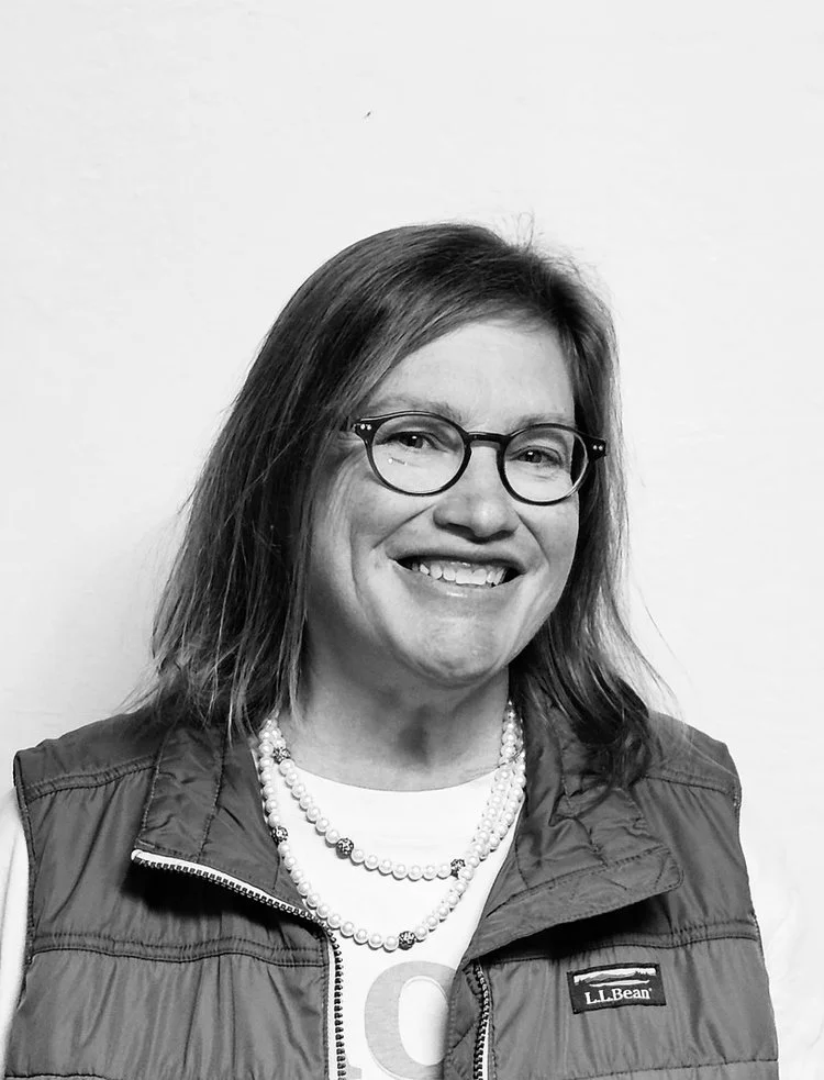 A smiling woman with shoulder length hair, wearing glasses and a sleeveless jacket and a necklace.