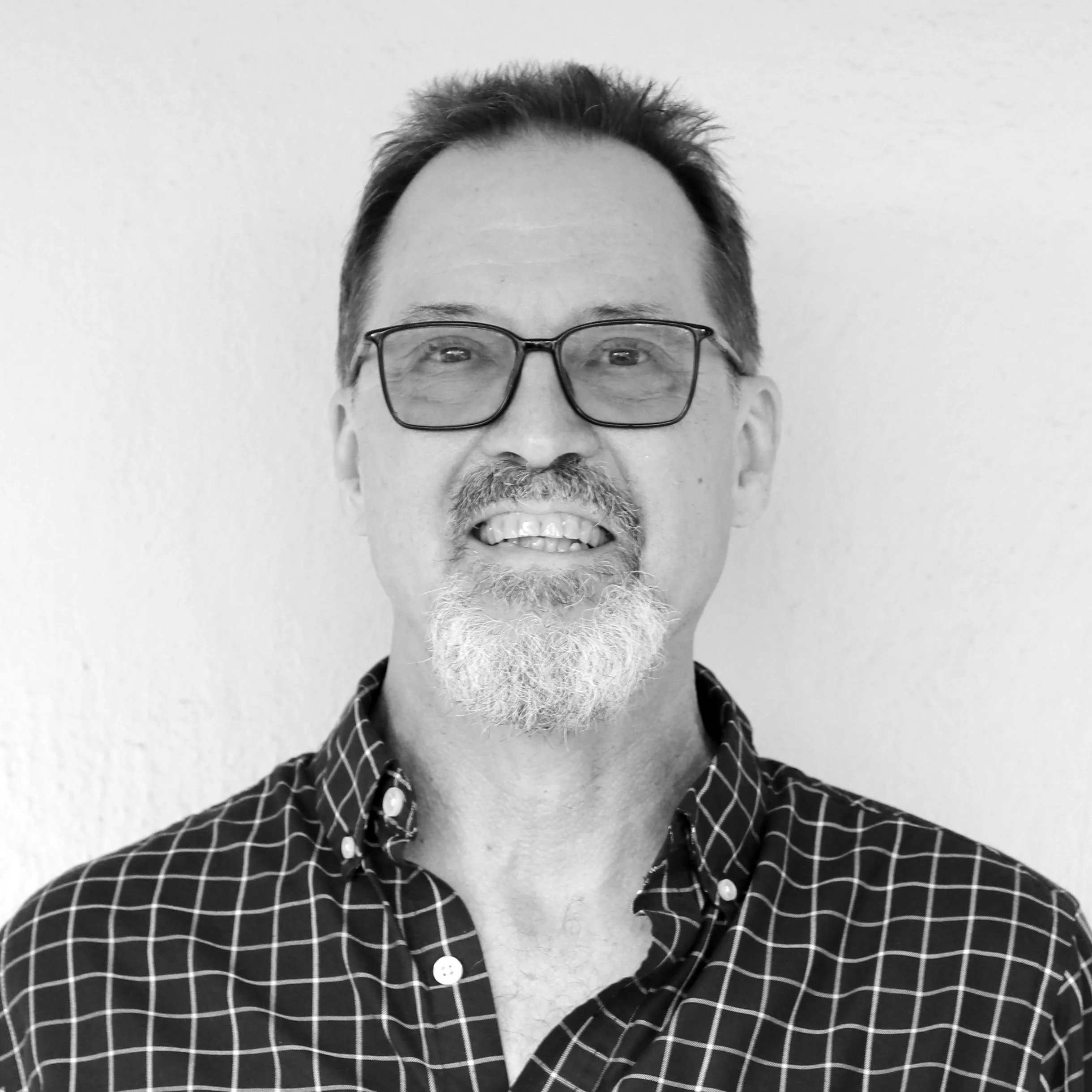 Black and white portrait of a middle-aged man with glasses, a goatee, and a checkered shirt, smiling.