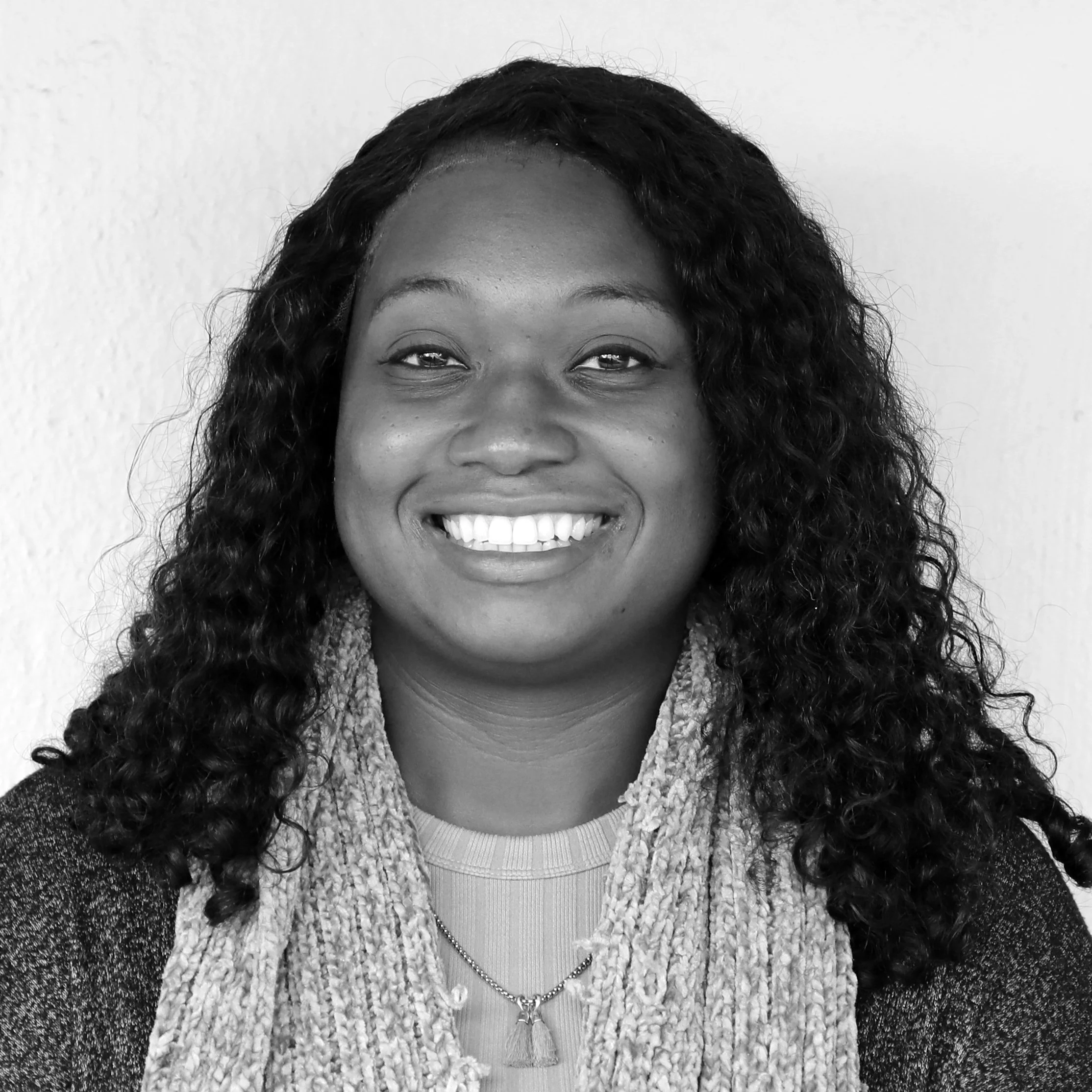 Black and white portrait of a smiling woman with long curly hair, wearing a textured scarf, a necklace, and a sweater, standing against a plain background.