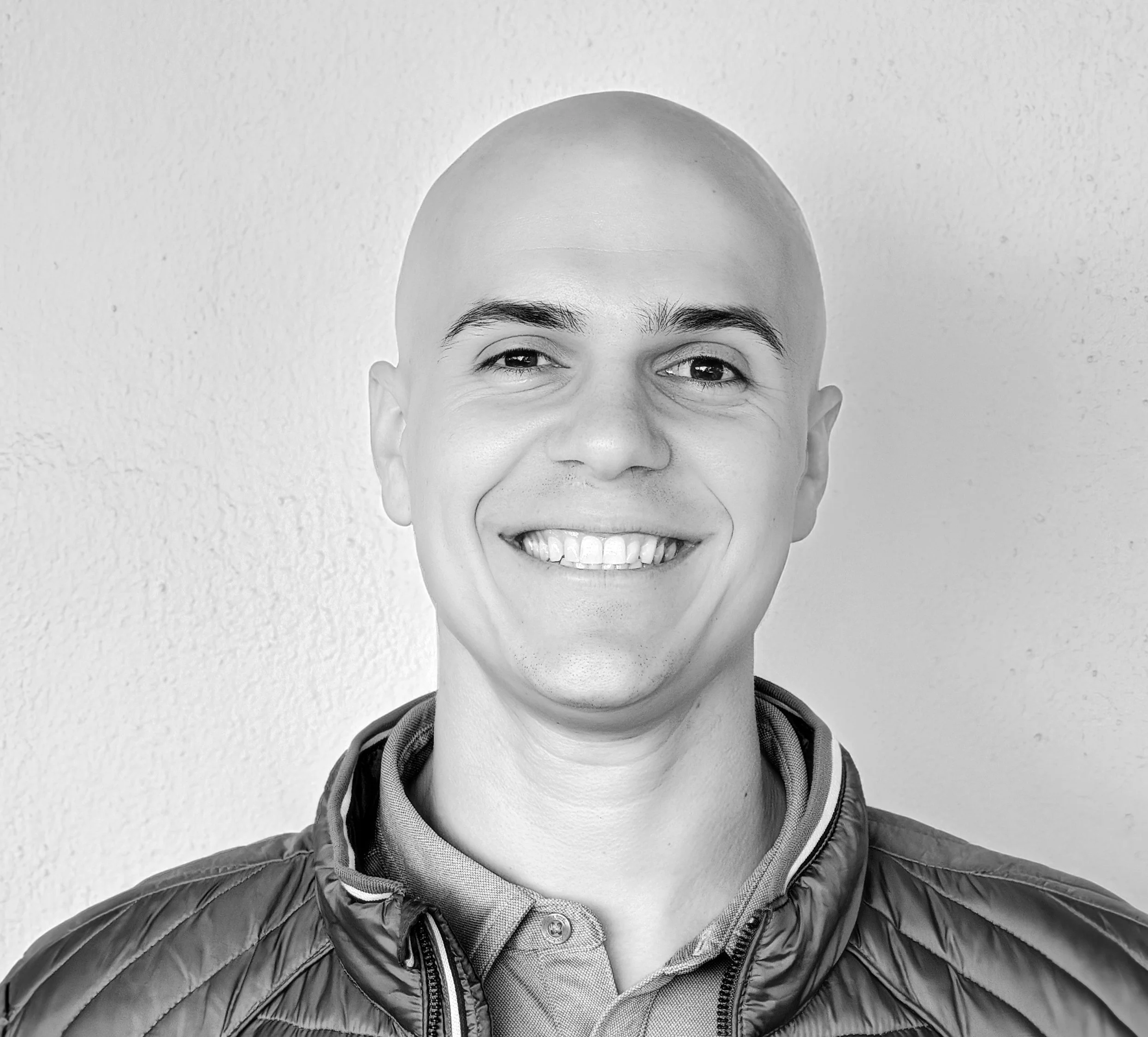 Black and white photo of a young man with short, dark hair, smiling, wearing a dark collared shirt, standing against a plain wall.