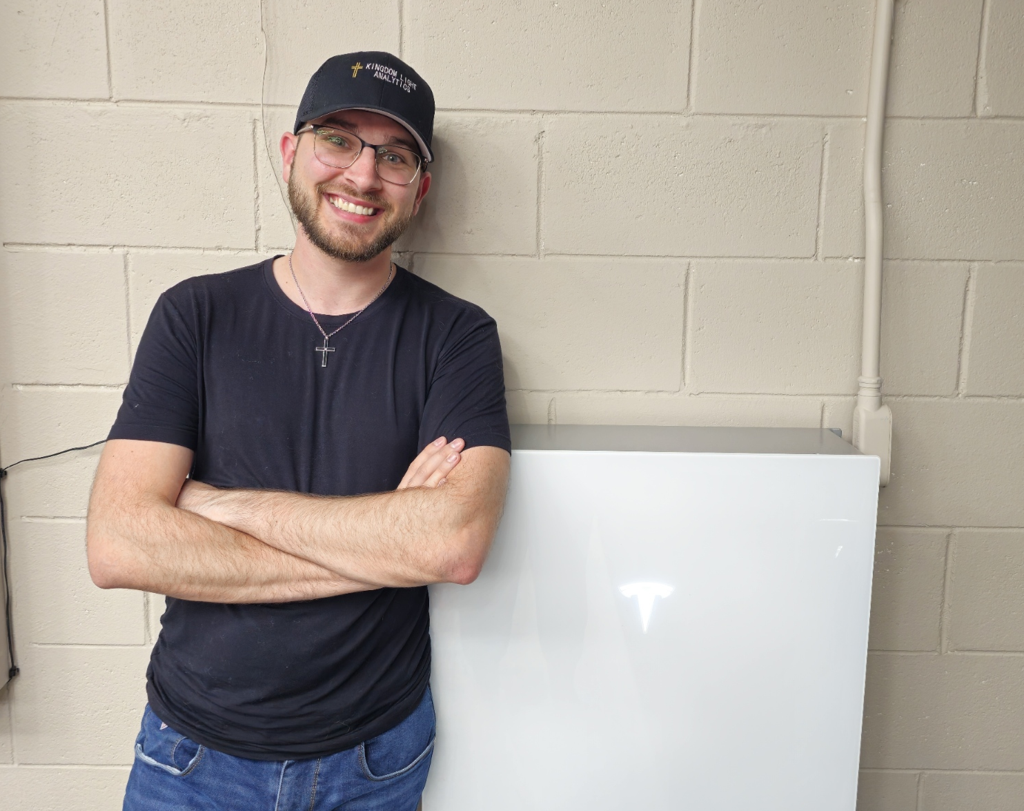 A man with glasses, a beard, and a cross necklace, smiling, standing with arms crossed next to a white box or container, in front of a beige brick wall.