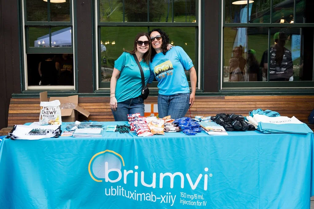 Two women standing behind a promotional table for a medical product called Bohrumvi, with various items displayed on the table, wearing blue shirts, outdoors with a building behind them.