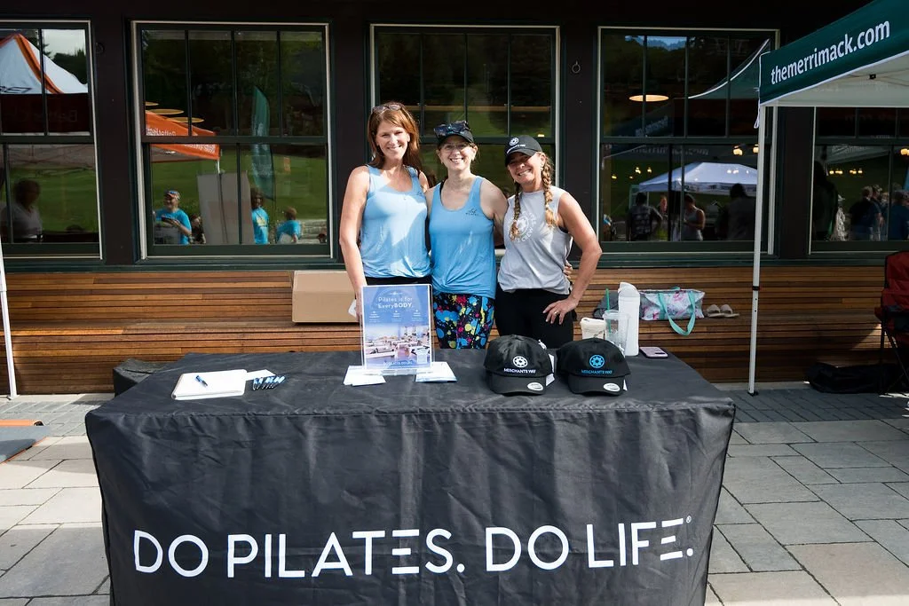 Three women standing behind a table with promotional items at an outdoor event. The tablecloth says 'Do Pilates. Do Life.' and has caps, papers, and other materials on it. Behind them are large windows reflecting the outdoor scene.