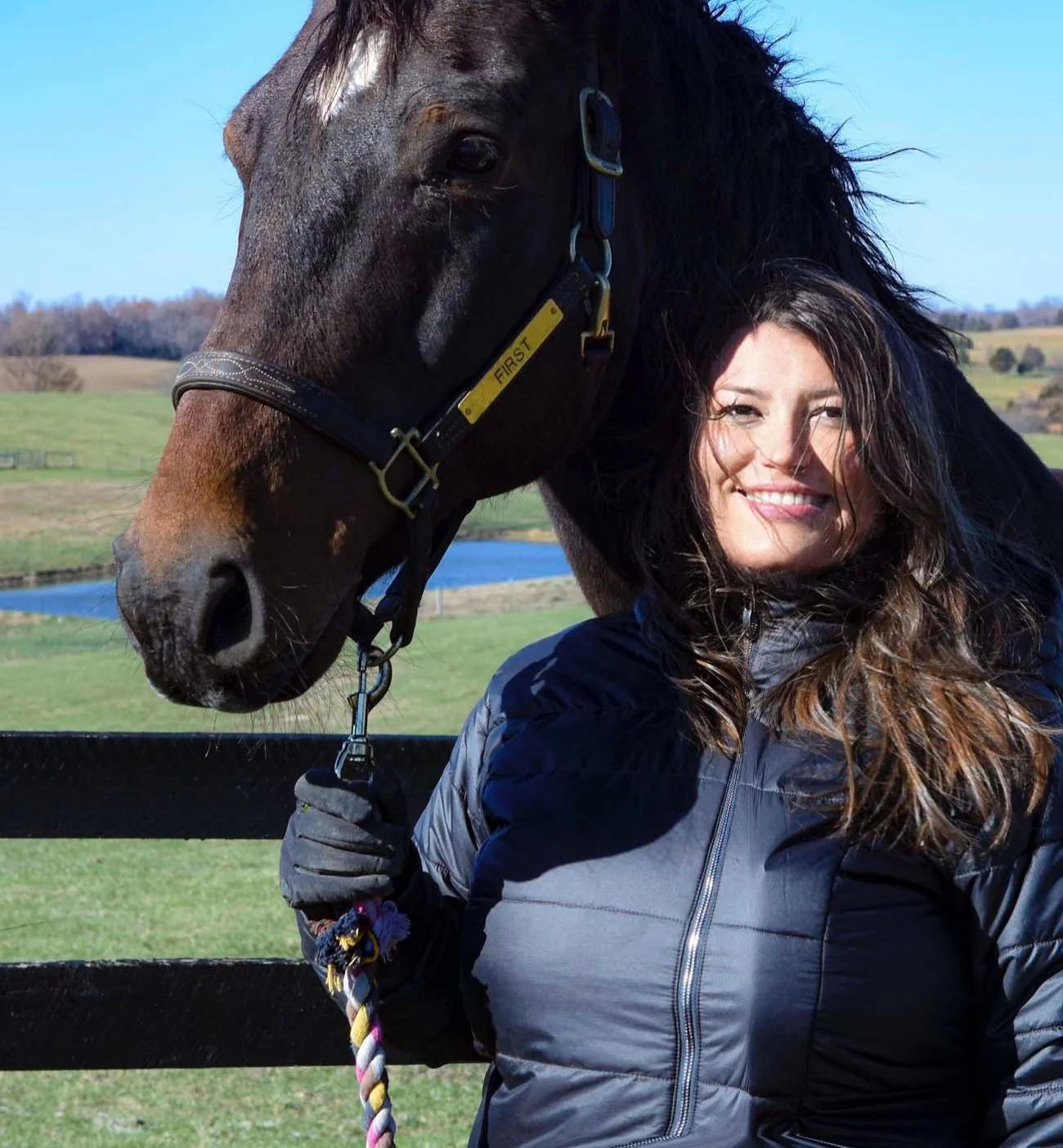 A woman standing next to a brown horse with black mane outdoors in a grassy field with a lake and hills in the background, sunny day.