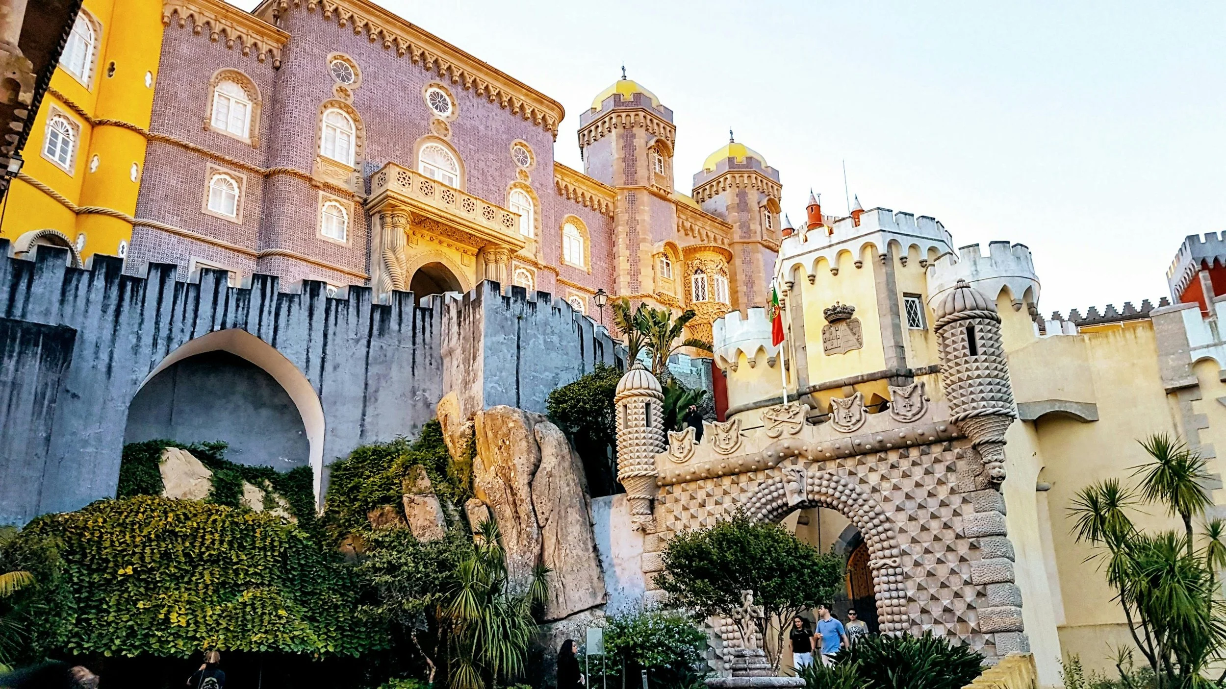 A colorful castle with pink and yellow walls, multiple towers, crenellated battlements, and decorative architectural details. There is a stone archway at the entrance, decorated with sculptures and textured patterns. Green plants and rocks are in the foreground with visitors walking nearby.