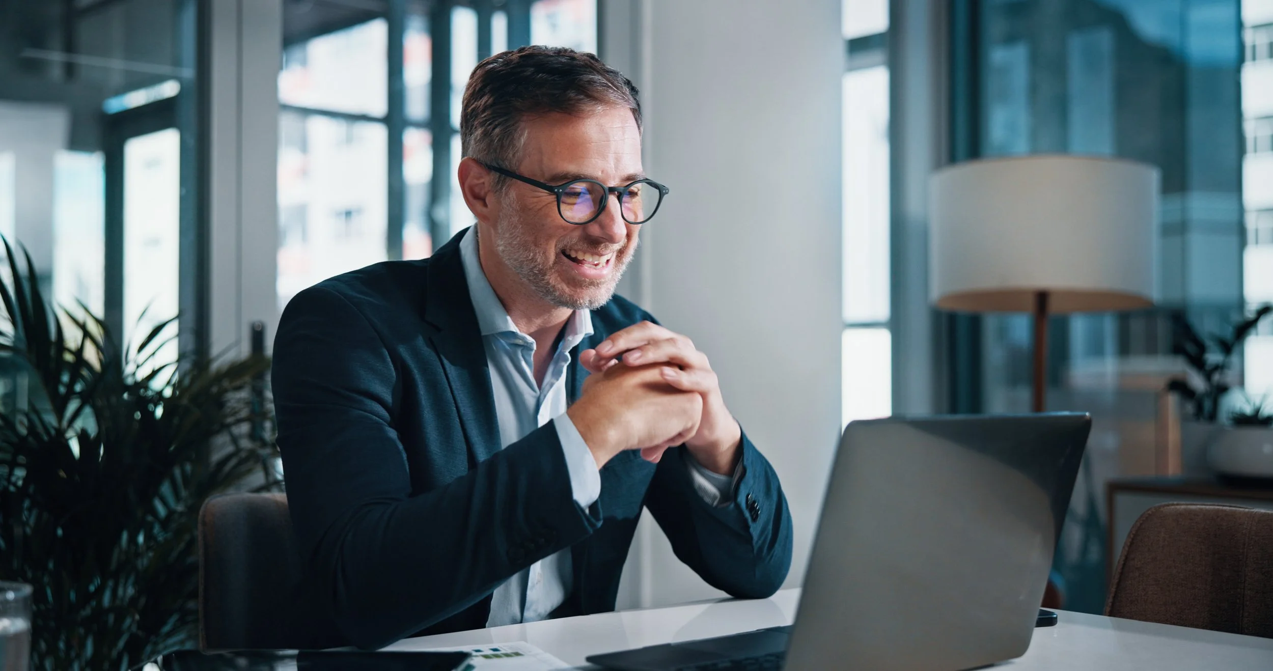 A middle-aged man with glasses smiling at a laptop in a modern office setting.