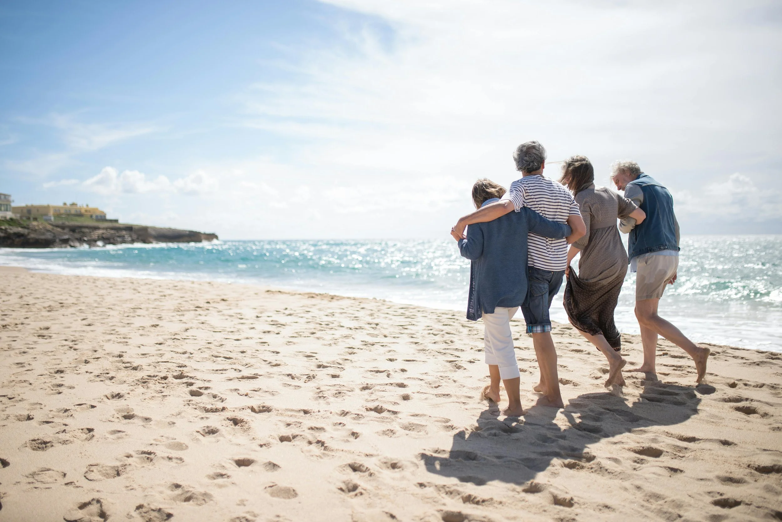 A group of four elderly people walking along a sandy beach near the ocean on a sunny day, with some of them embracing each other.