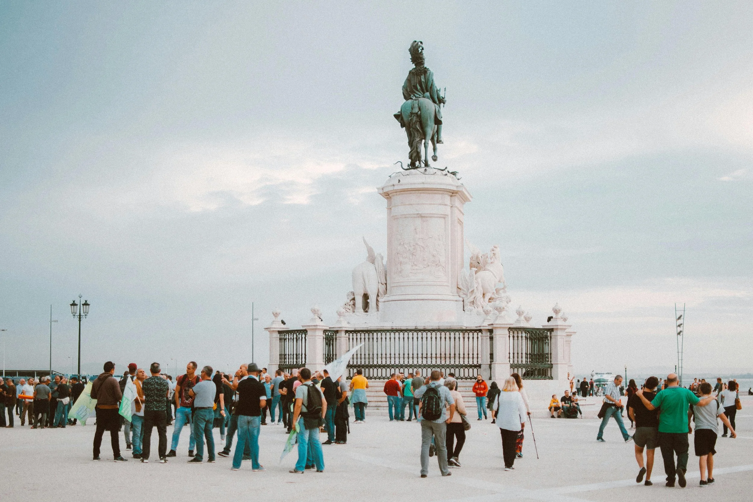 Crowd of people gathered around a large monument with an equestrian statue on top in an open plaza.