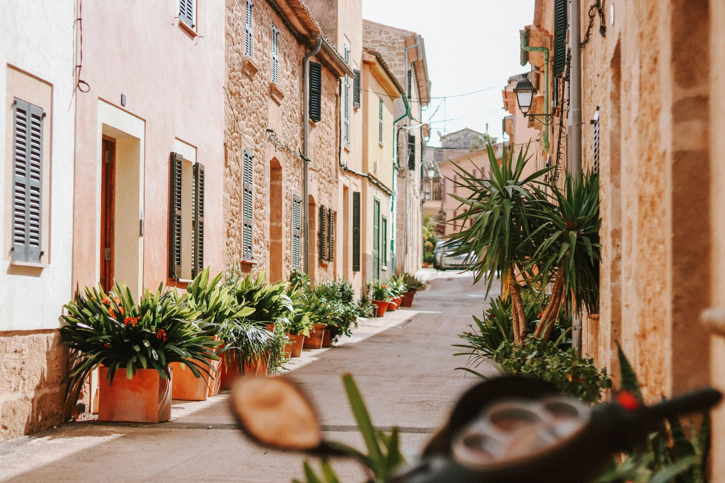A narrow street in a European village with pastel-colored buildings, potted plants, and a parked car at the end of the street.