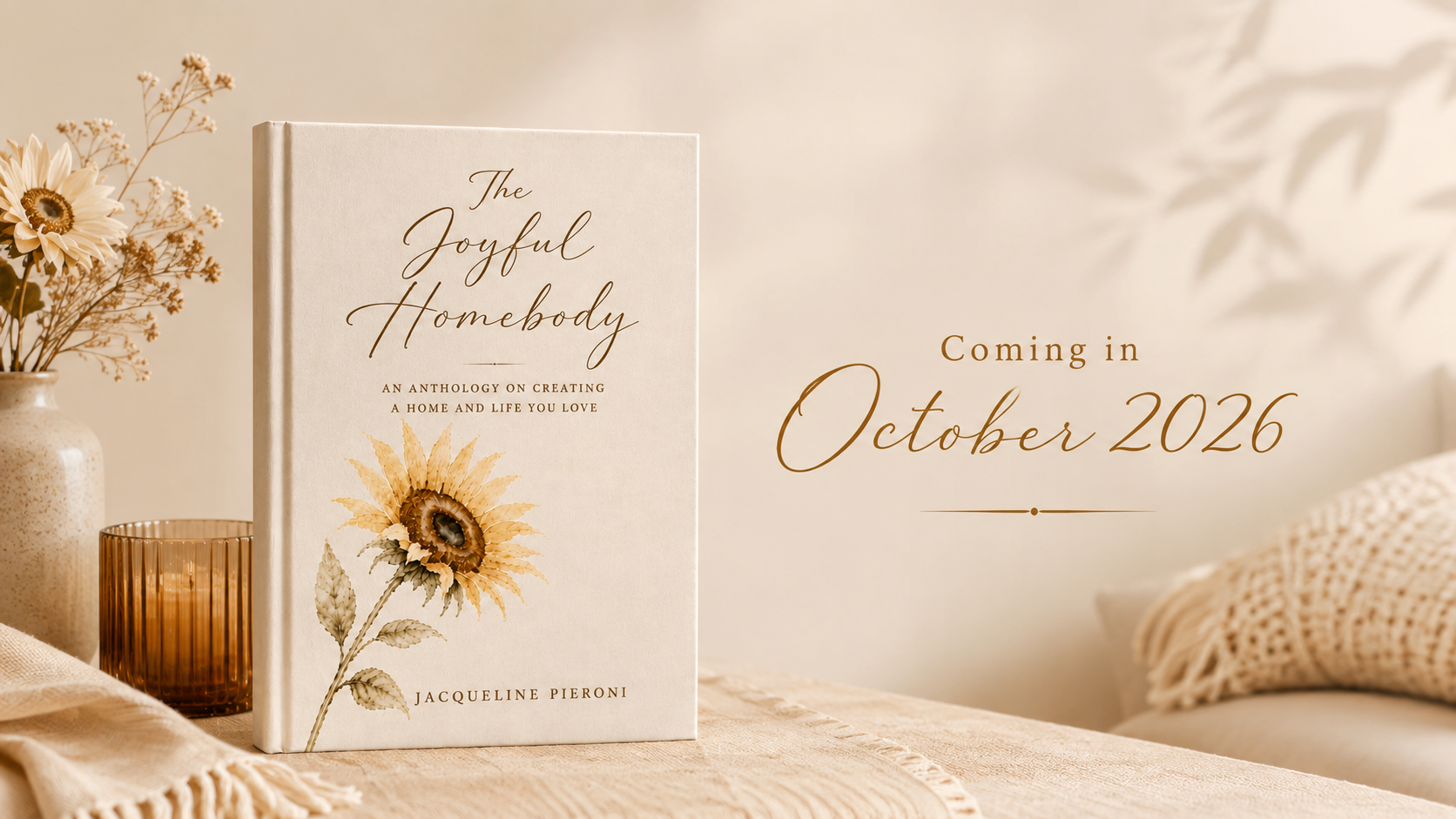 A book titled "The Joyful Homebody" by Jacqueline Pieroni resting on a beige textured surface, next to a small vase with dried flowers, and a brown glass candle holder, with a blurred background of a cozy room and text indicating a new release in October 2026.