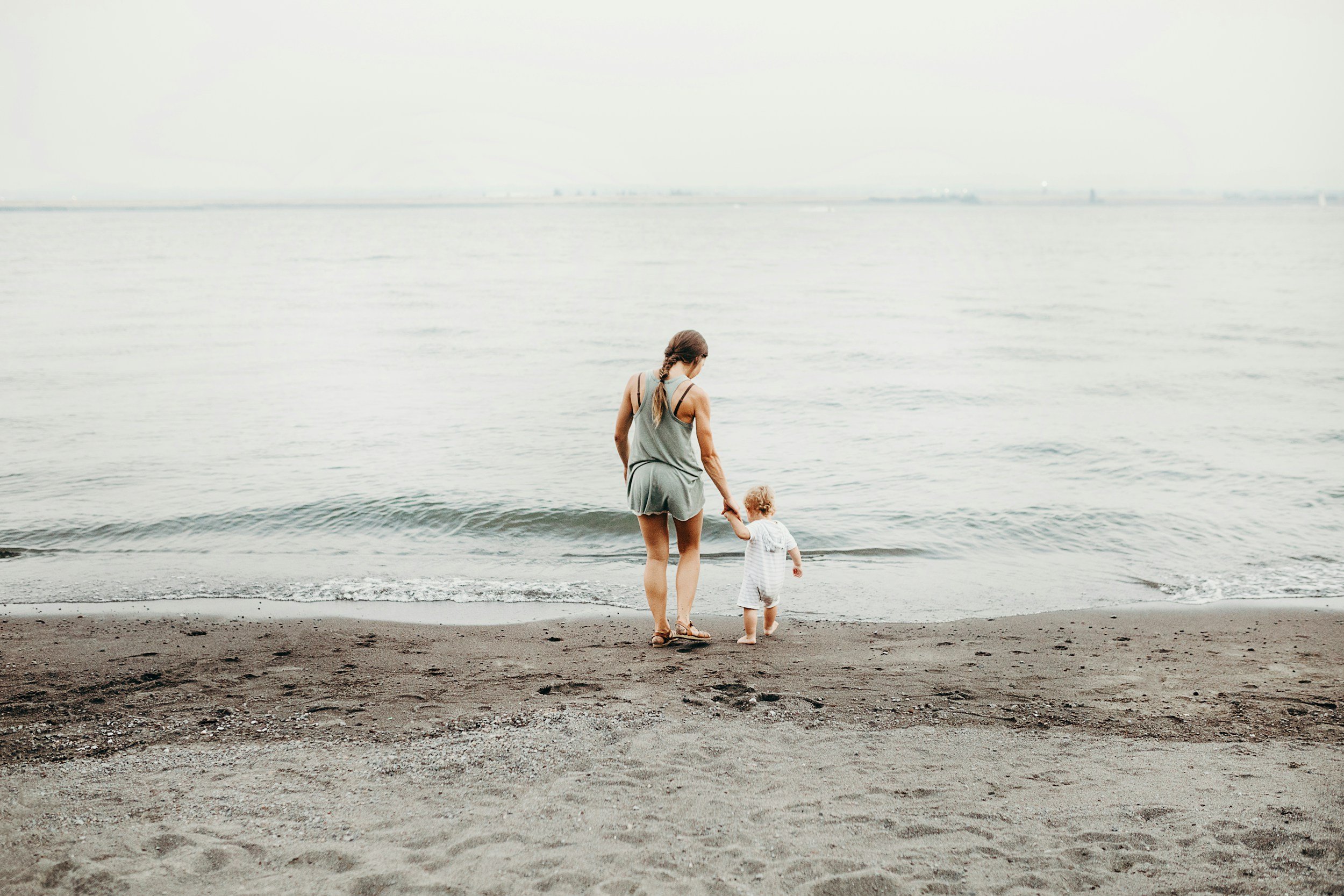 A woman and a young child walking hand-in-hand along the beach shoreline, facing the water.