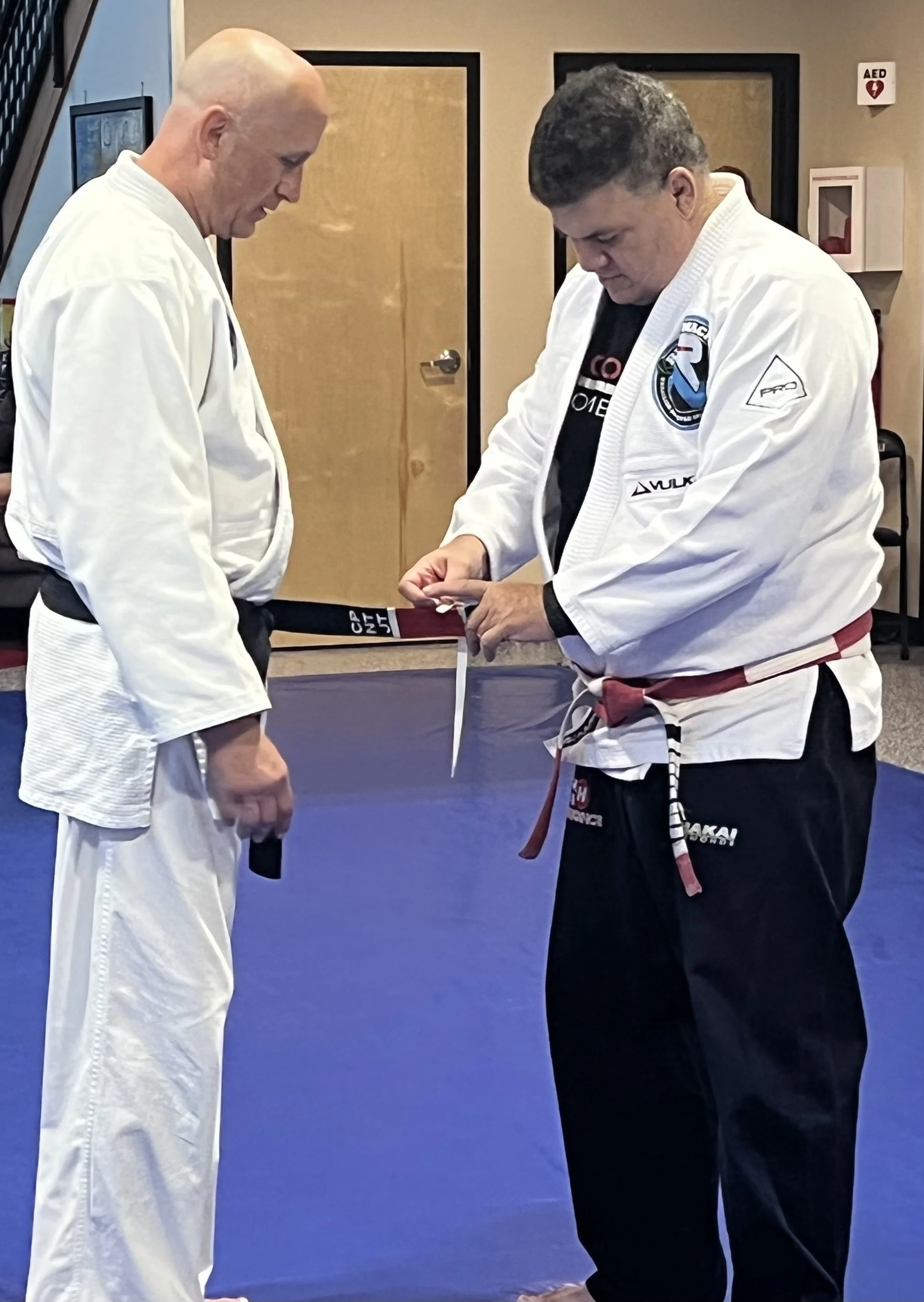 Two men in martial arts uniforms, or gi, standing on a blue mat. One man with a black belt is tying his belt while the other man looks on. They are indoors, possibly in a martial arts training facility.