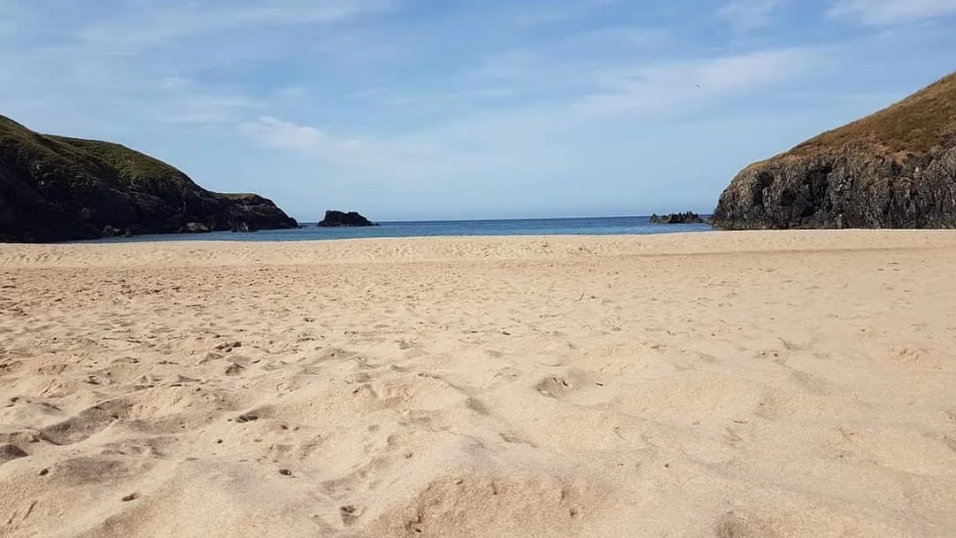 An empty sandy beach surrounded by rocky cliffs under a clear blue sky.