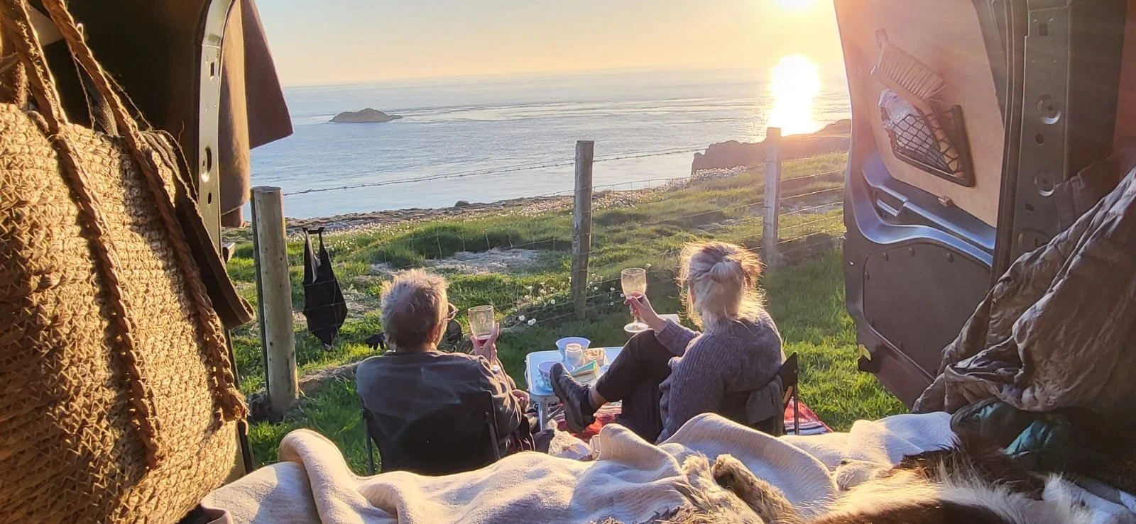 View from inside a camper van showing two people sitting and enjoying the sunset by the beach, with glasses of wine on a small table.