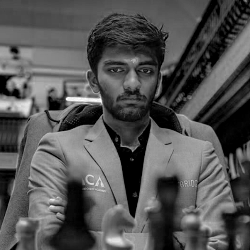 Black and white photo of Gukesh Dommaraju  with short dark hair and a beard, wearing a blazer and sitting at a table with chess pieces in the foreground.