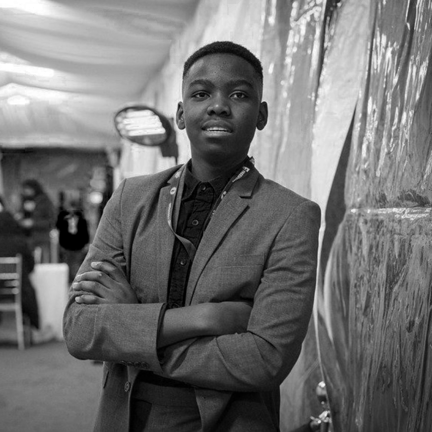 Tani Adewumi in a suit standing with crossed arms, looking at the camera, in an indoor setting with blurred background.