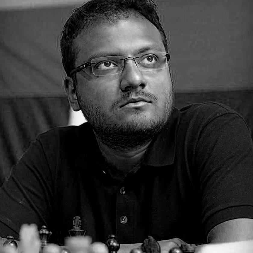 Black and white photo of Sagar Shah with short dark hair, glasses, and a beard, looking thoughtful, sitting at a table with chess pieces.