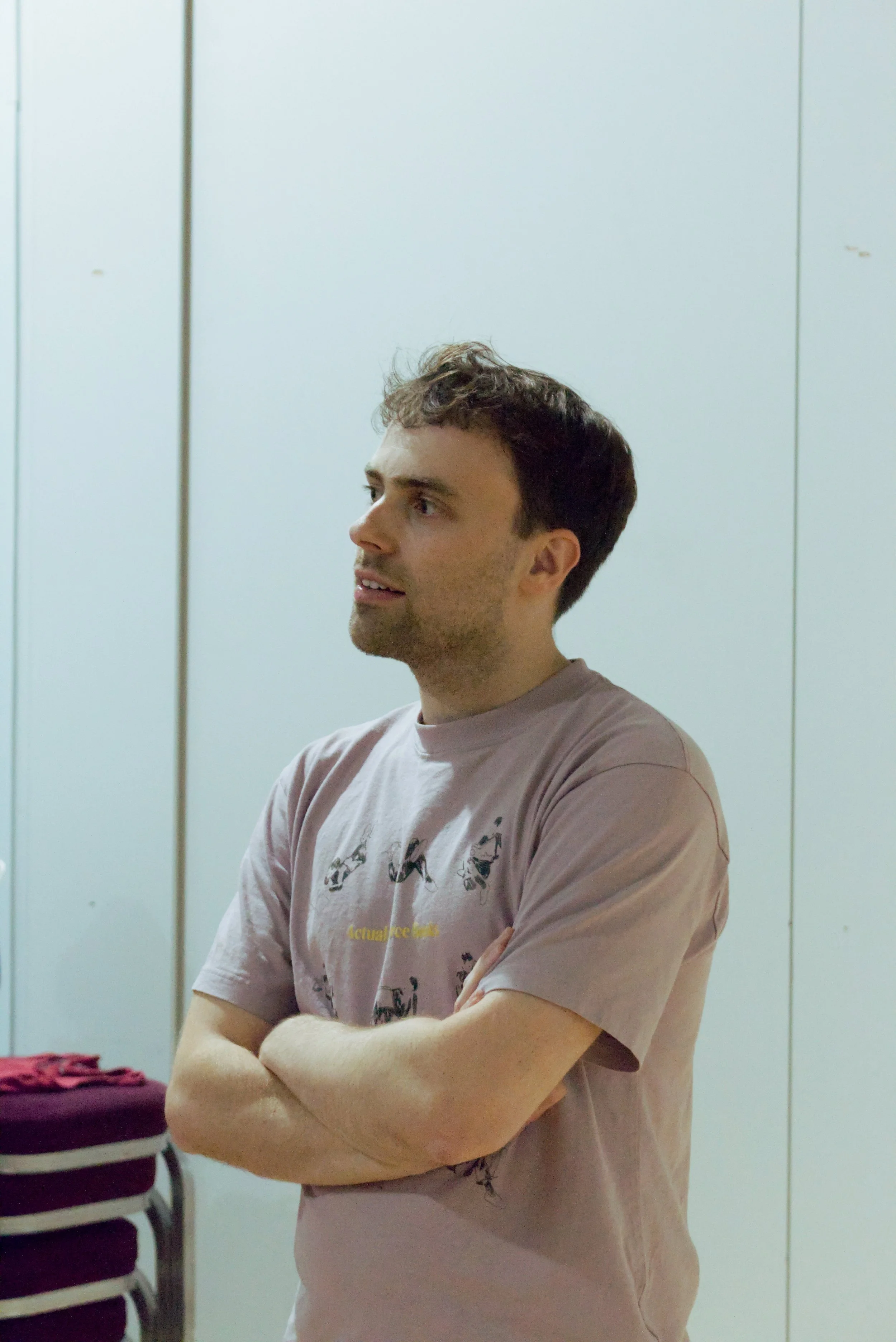 A young man with short brown hair and light skin wearing a light pink T-shirt with a graphic print, standing with arms crossed in front of a white wall with metal paneling.