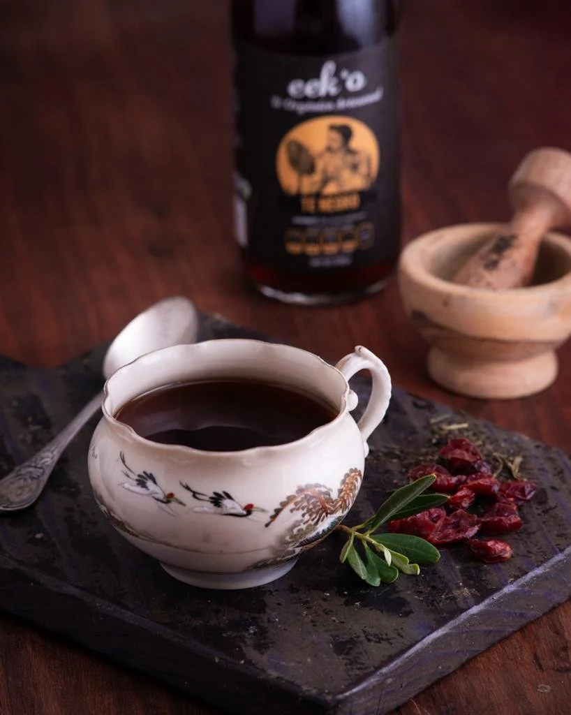 Taza de té negro en porcelana con ilustraciones, cucharita, mortero de madera y botella de té negro al fondo, sobre superficie de madera.