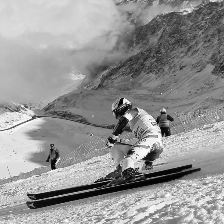 Praticante de esqui descendo uma encosta de neve em uma montanha, com outros praticantes ao fundo, céu nublado e montanhas ao redor.