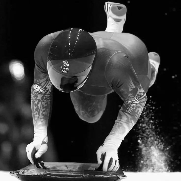 Atleta de esportes aquáticos em posição de start, usando capacete, luvas e traje de competição, em uma piscina artística, com uma iluminação de fundo.