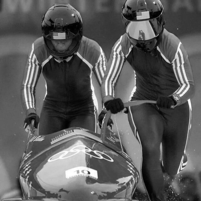 Duas pessoas com roupas esportivas e capacetes jogando tritã em uma embarcação de corrida.