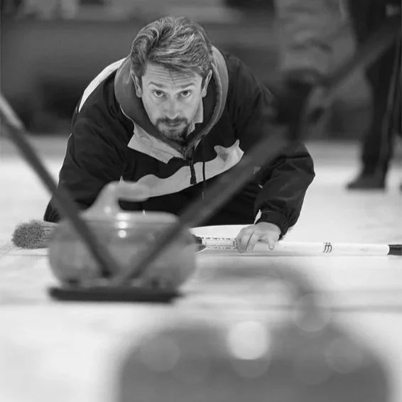 Homem com cabelo e barba, usando jaqueta, olhando para a câmera enquanto joga boliche.