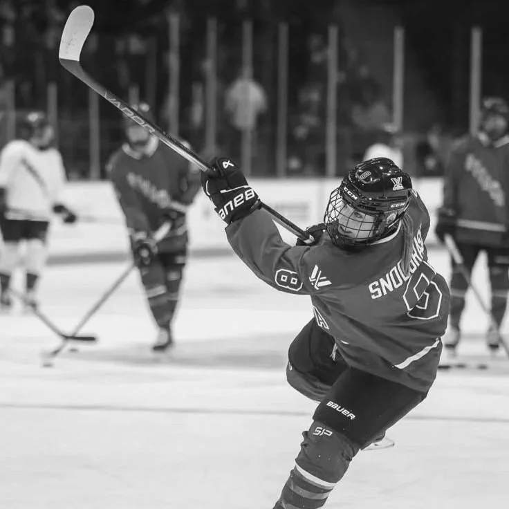 Jogador de hóquei no gelo com capacete e equipamento, segurando um bastão, em uma pista de gelo com outros jogadores ao fundo.