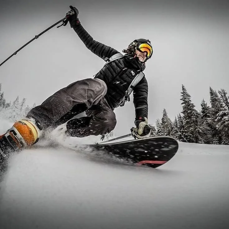 Pessoa praticando snowboard em uma pista de neve, vestindo roupas de inverno e usando capacete e óculos de sol, com árvores na paisagem ao fundo.