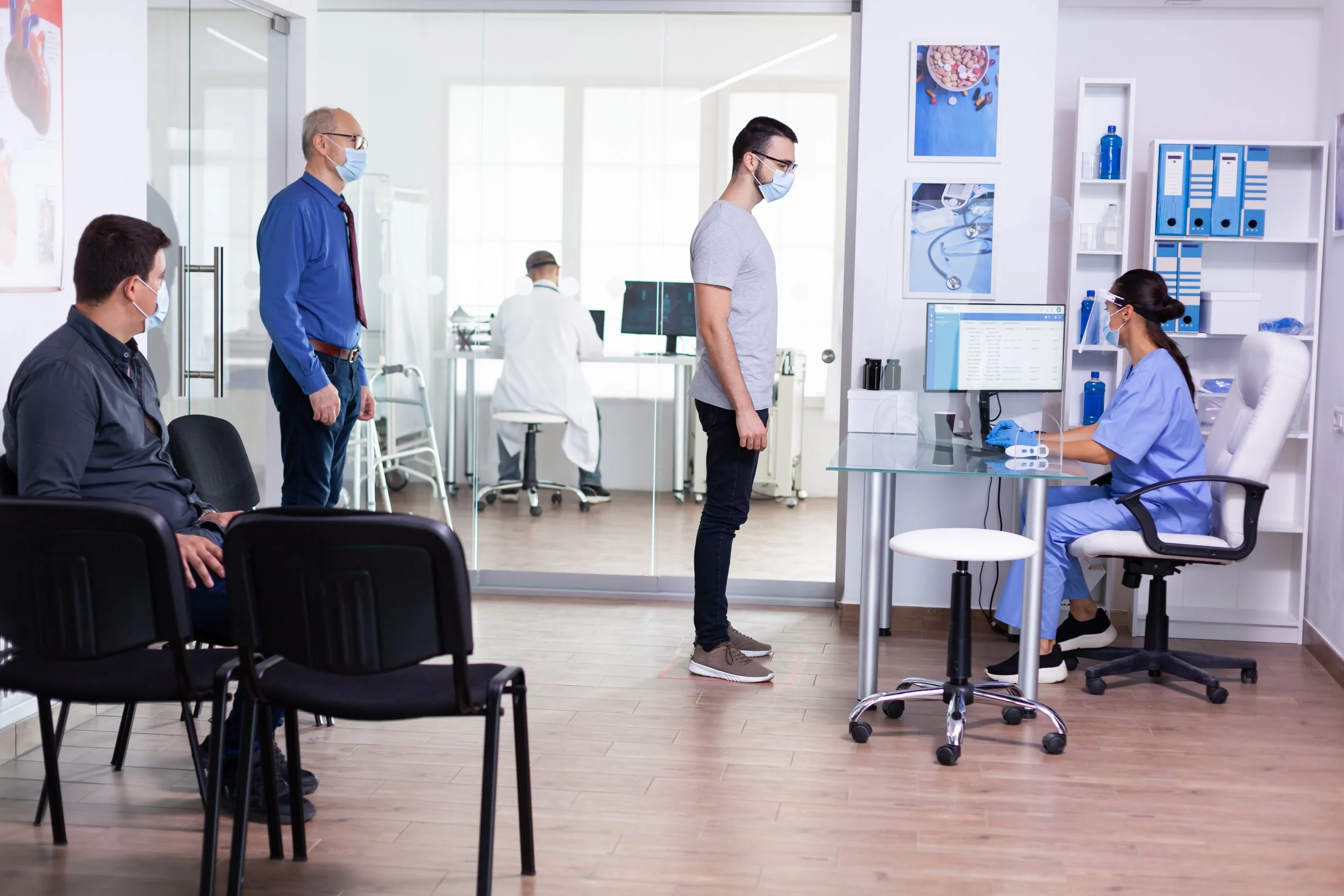 A person standing in a hospital or clinic waiting room while maintaining social distancing, with three other people and a medical professional working at a desk, all wearing face masks.