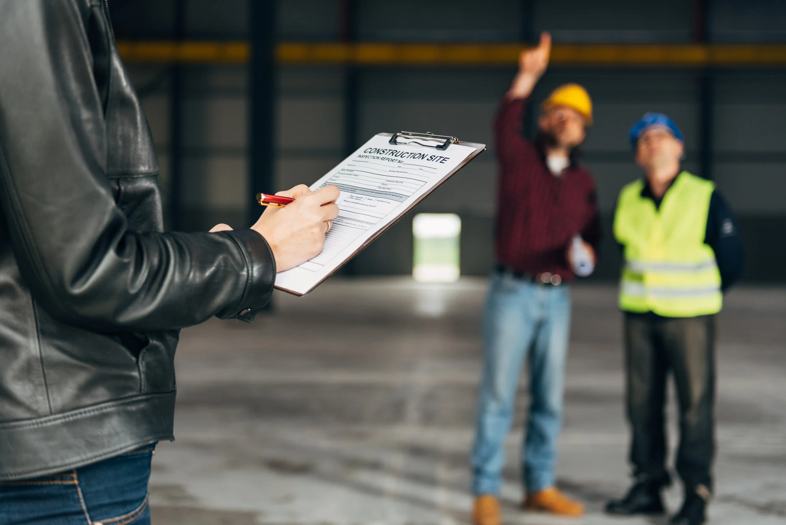 Person with a leather jacket holding a clipboard with a construction site inspection report, two workers in safety helmets and vests raising hands and talking inside a warehouse or industrial building.