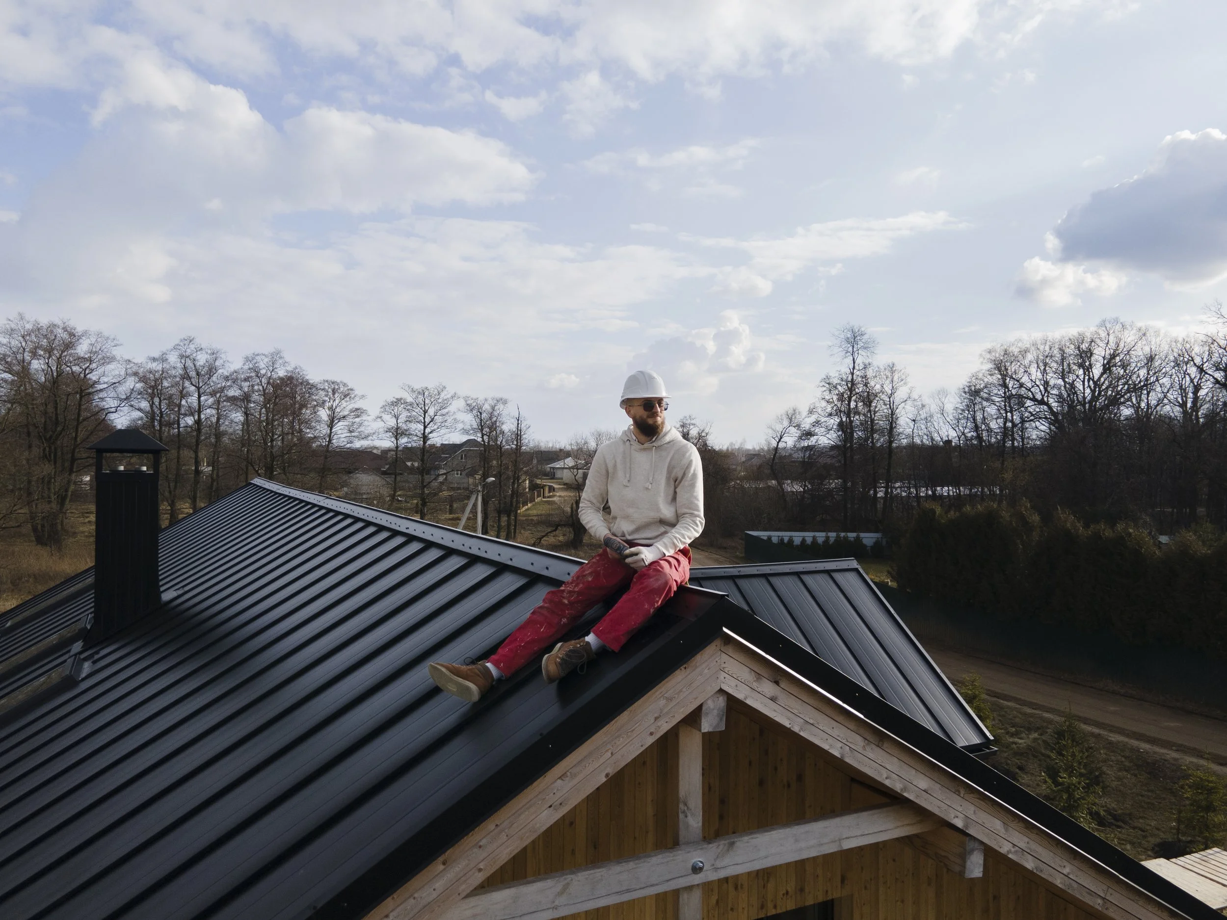 A man wearing safety gear, including a helmet and sunglasses, sitting on a roof with a metal surface, surrounded by trees and houses in the background under a cloudy sky.