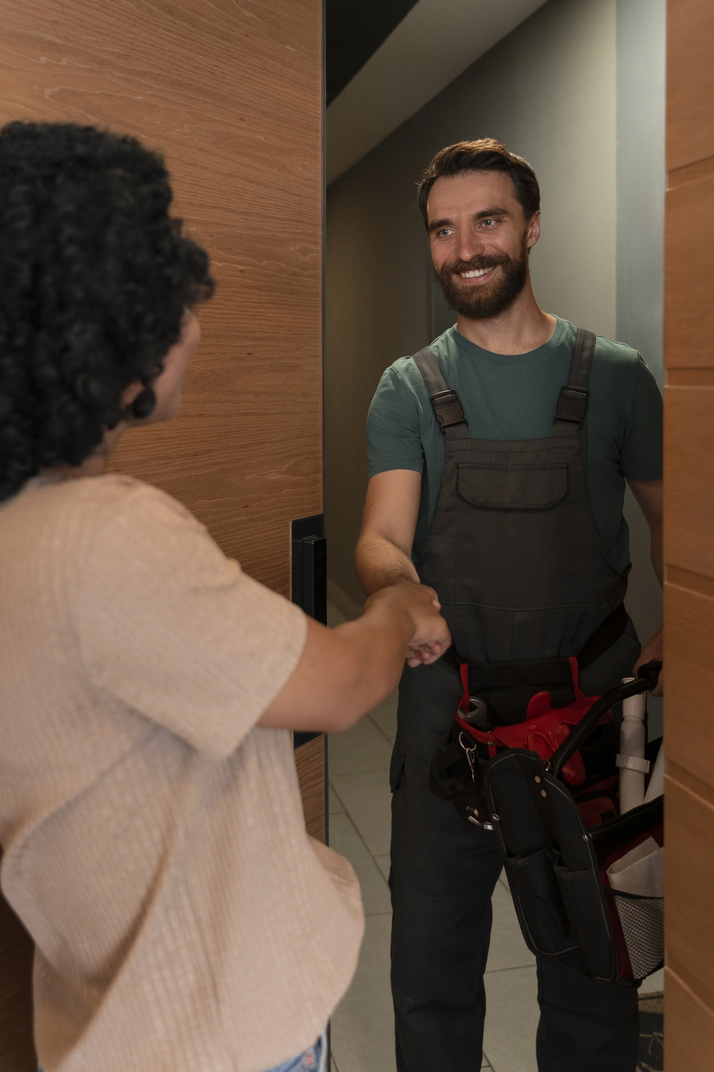 A man in a green t-shirt and work overalls shakes hands with a woman at a doorway.