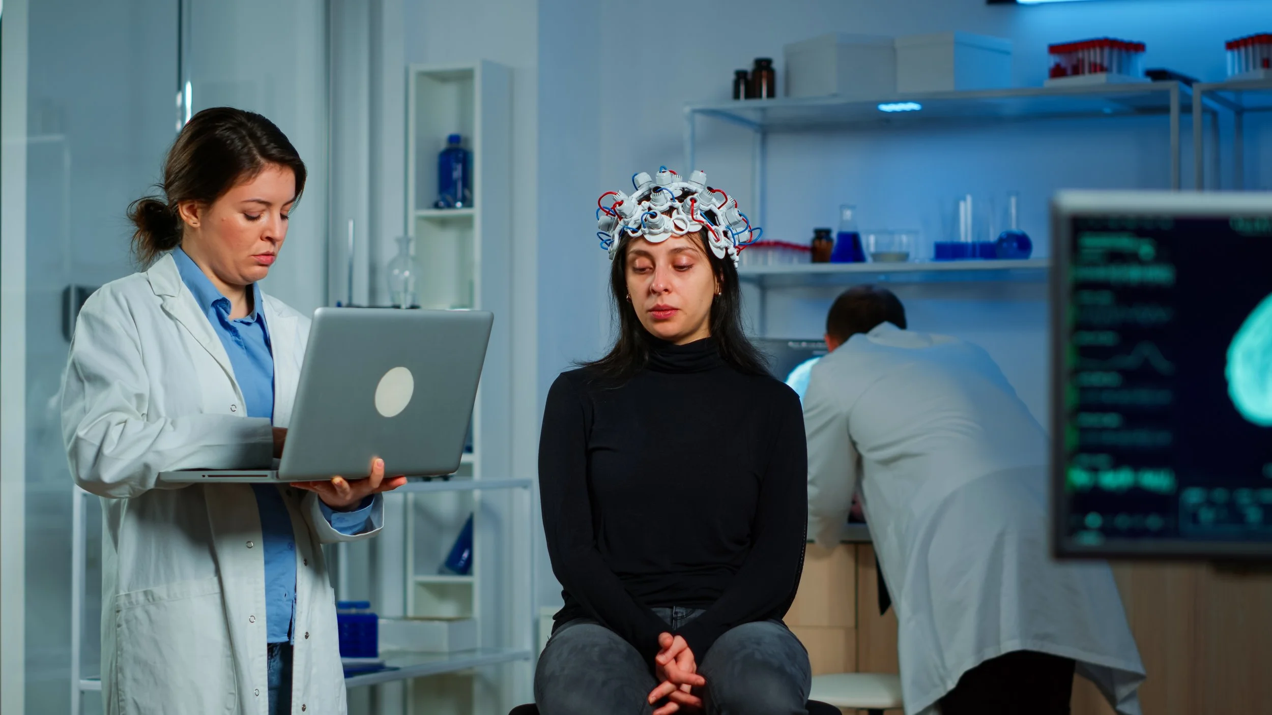A woman in a black shirt sits with her eyes closed and a cap made of wires and electronic gadgets on her head, while a female doctor in a lab coat holds a laptop and looks at it in a laboratory setting with medical equipment and another person in a white coat working in the background.