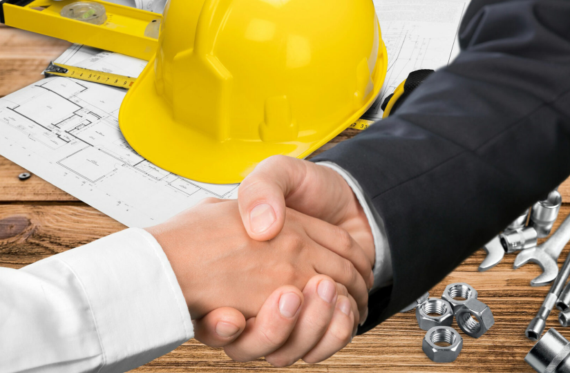 Two people shaking hands over a wooden table with architecture plans, a yellow safety helmet, and mechanical tools and nuts nearby.
