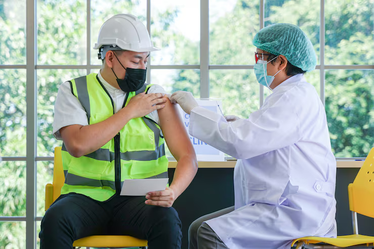 A healthcare professional in a white coat, blue surgical cap, and face mask administering a vaccination to a man in construction attire, including a white helmet, high-visibility vest, and black mask, in a well-lit room with large windows revealing green outdoor scenery.