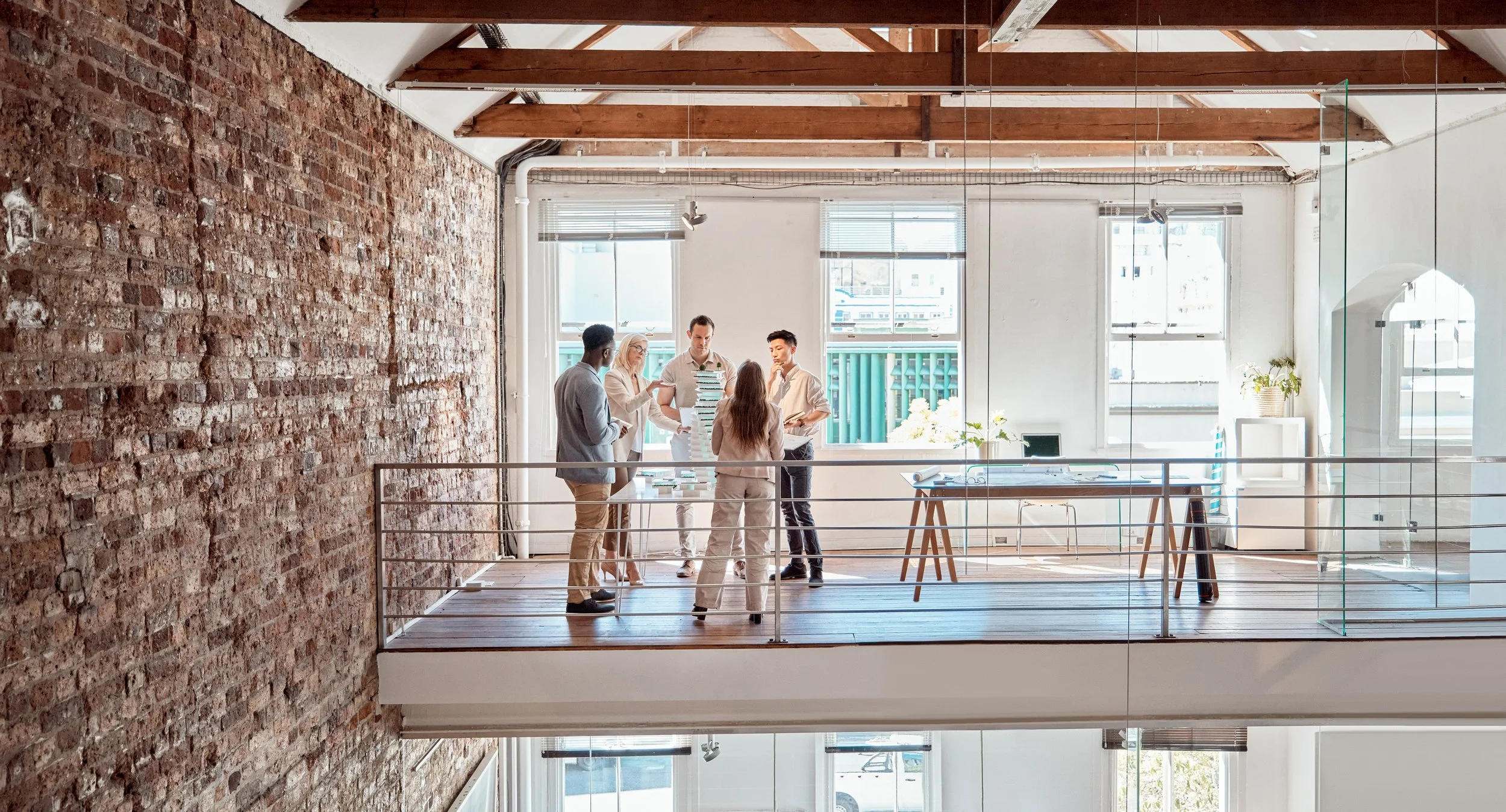 A group of five business professionals standing around a table on an open loft office space, engaging in a discussion, with a brick wall on the left and large windows in the background.