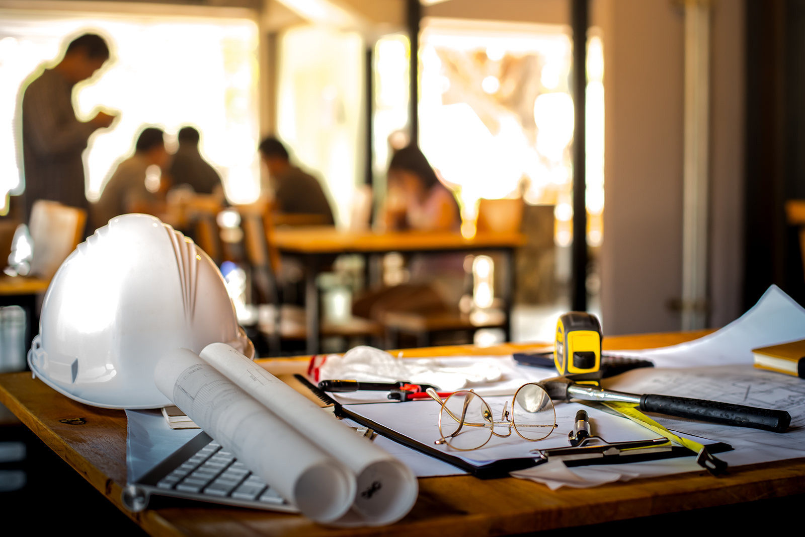 Construction or architectural workspace with a white hard hat, blueprints, a tape measure, glasses, and various tools on a wooden table, with people working in the background.