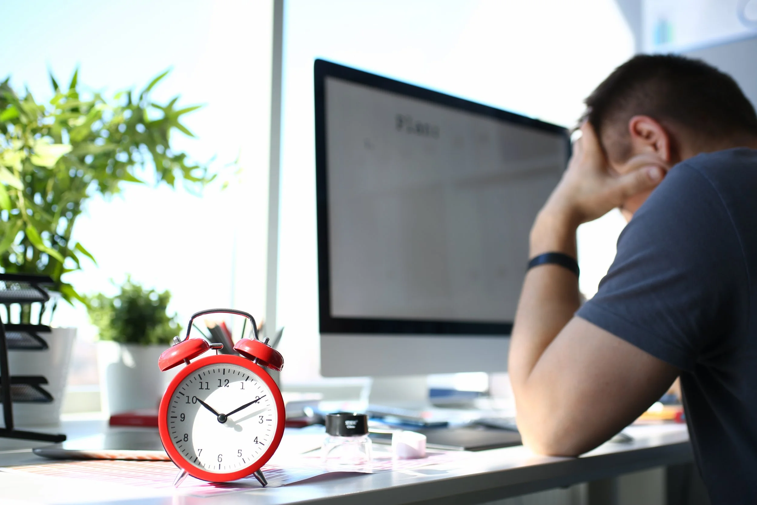 A red alarm clock showing 10:10 sitting on a cluttered desk with a person in a dark shirt resting their head on their hand in front of a computer monitor, in a brightly lit room with green plants nearby.
