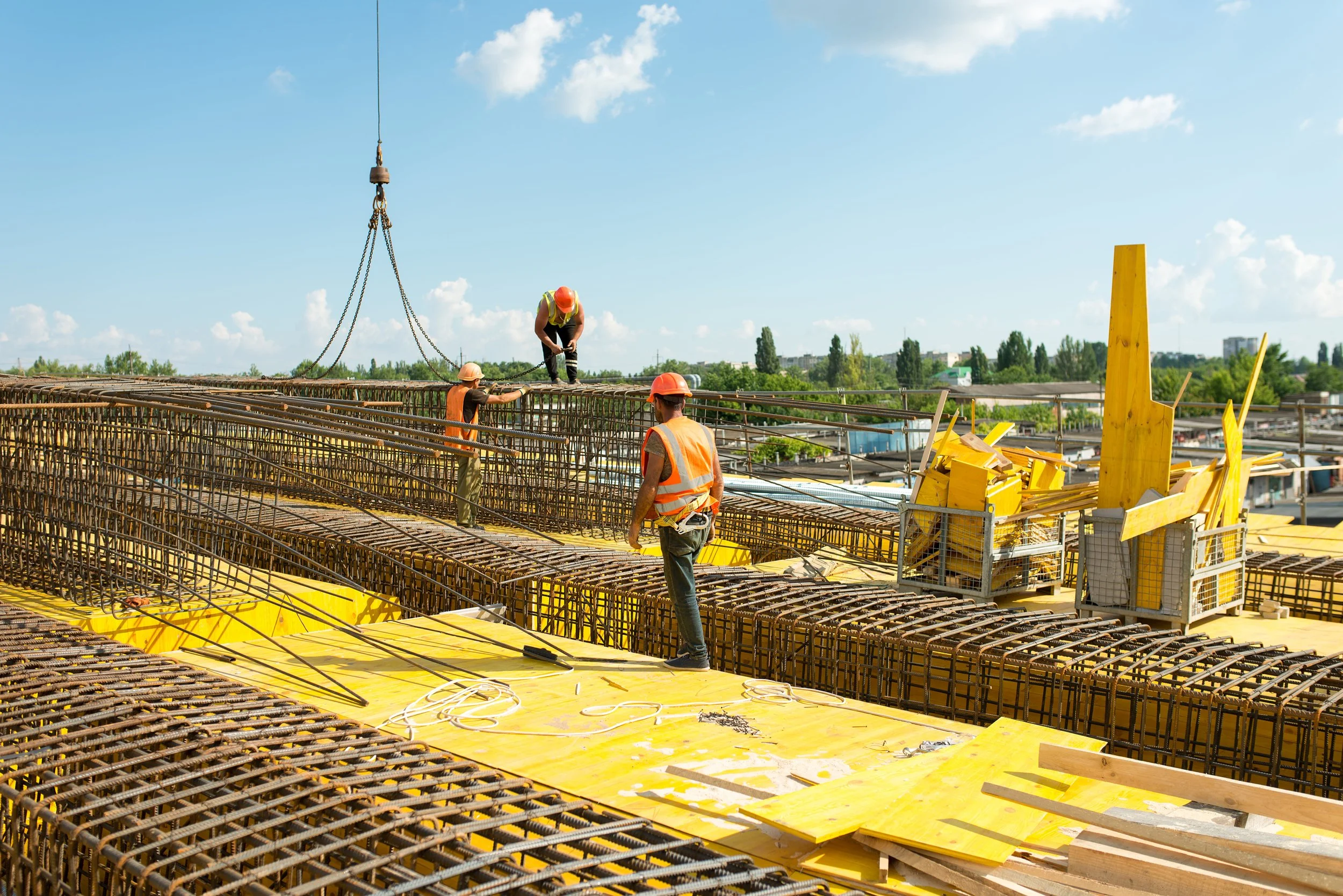 Construction workers on a building site working on reinforcement steel bars with tools and yellow safety equipment, under a blue sky with scattered clouds.