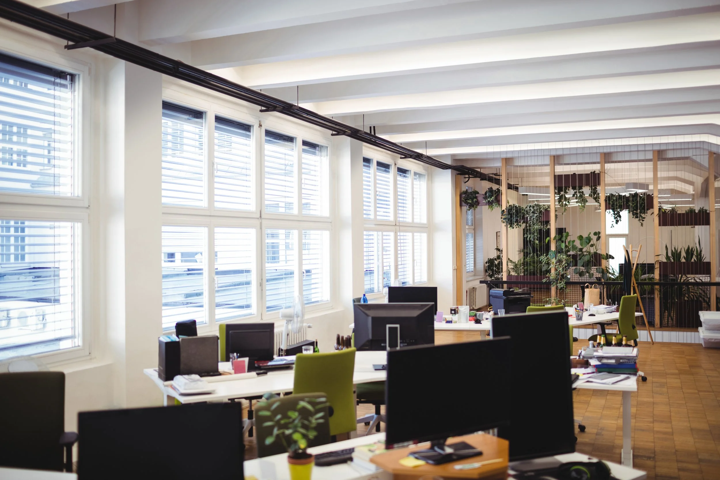 Open office space with multiple desks, computers, chairs, and large windows with blinds, decorated with plants and wooden dividers.