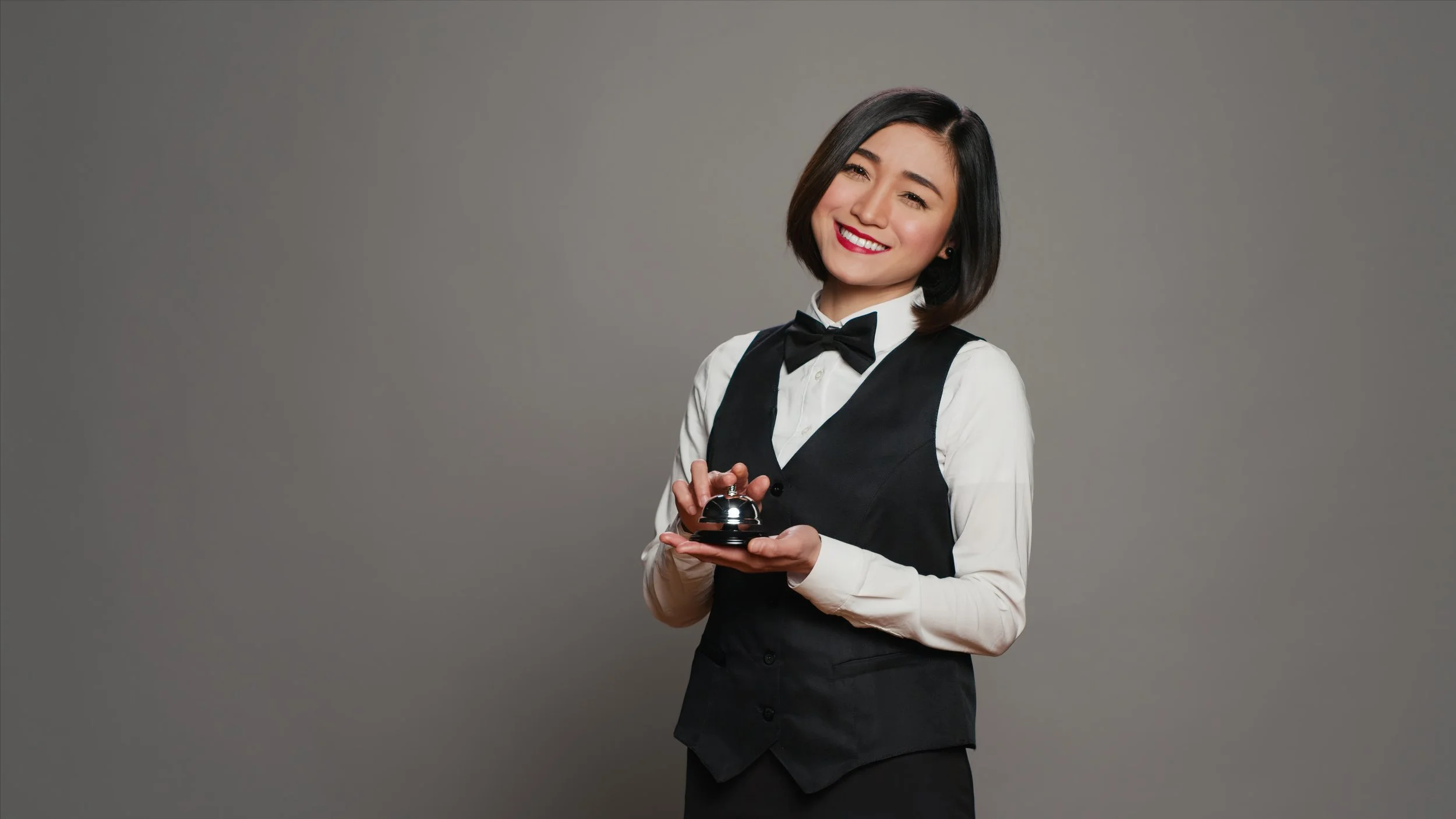 A young woman dressed as a hotel or service industry employee, wearing a white shirt, black vest, and bow tie, holding a service bell and smiling.