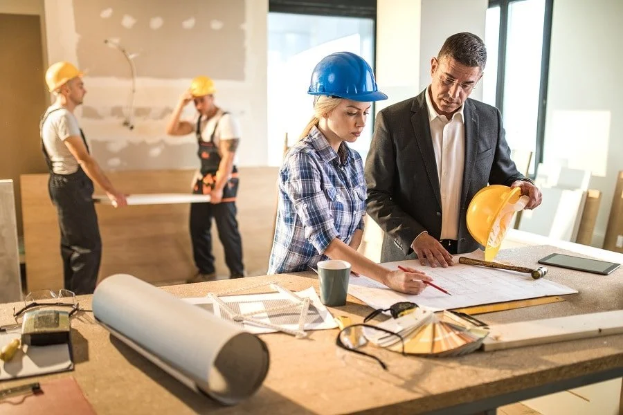 Interior of an office or construction site with five people; two workers in safety helmets in the background, and a woman and man in the foreground reviewing blueprints, with the woman wearing a blue helmet and the man holding a yellow helmet.