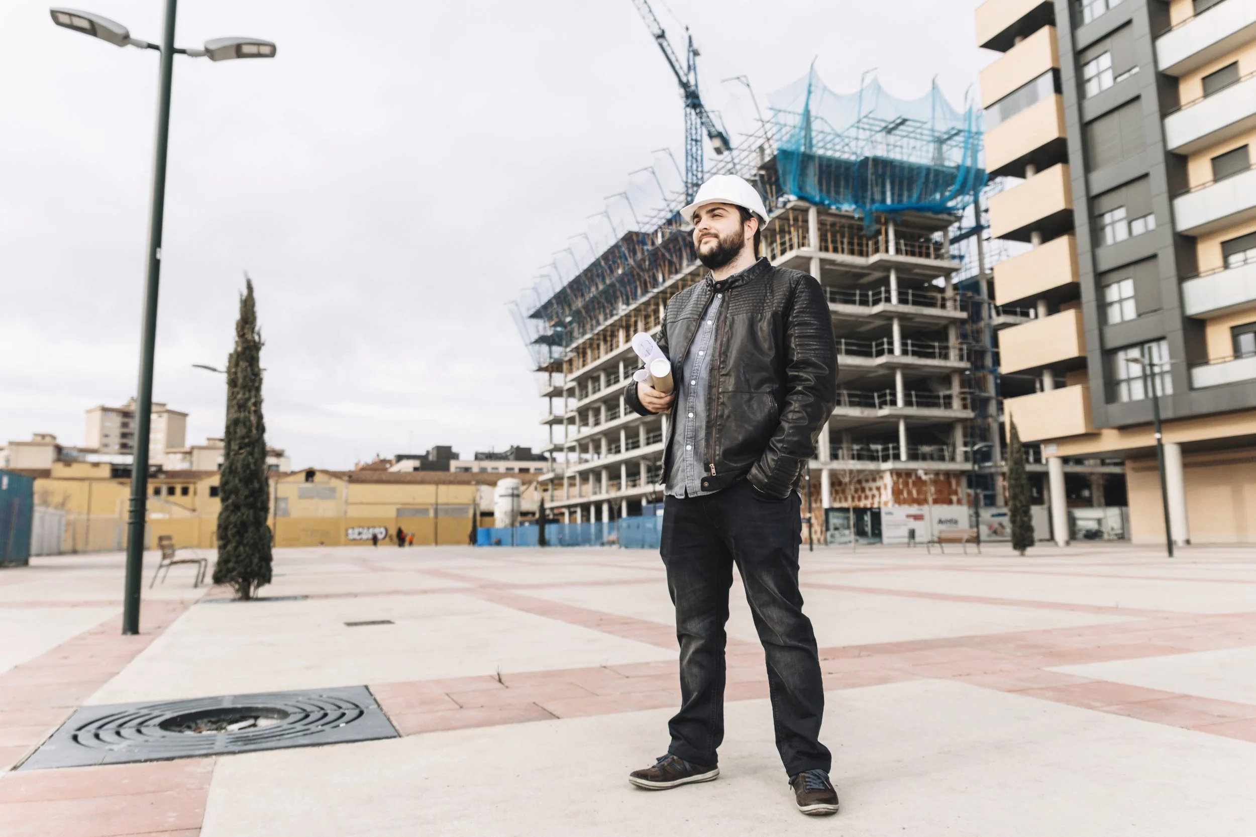 A man in a construction helmet and leather jacket holds rolled-up papers and stands in an urban area with partly constructed buildings, trees, benches, and modern apartment buildings in the background.
